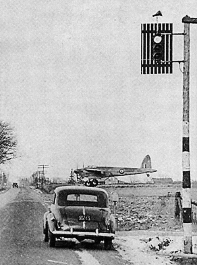 A black and white photo of a car stopped at a traffic light, in front of a runway and a de Havilland Mosquito aircraft.