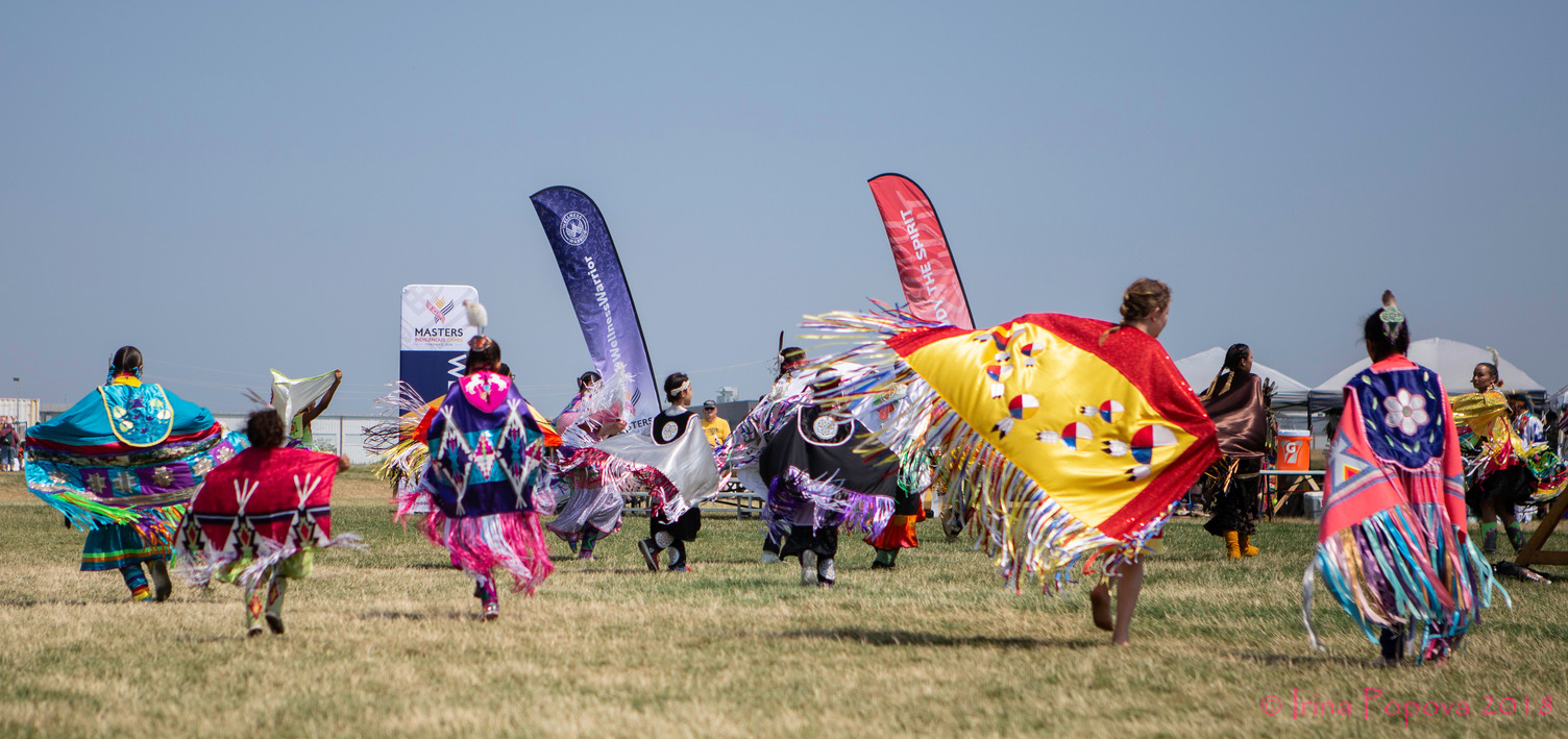 Indigenous dancers on a field, wearing colourful shawls.