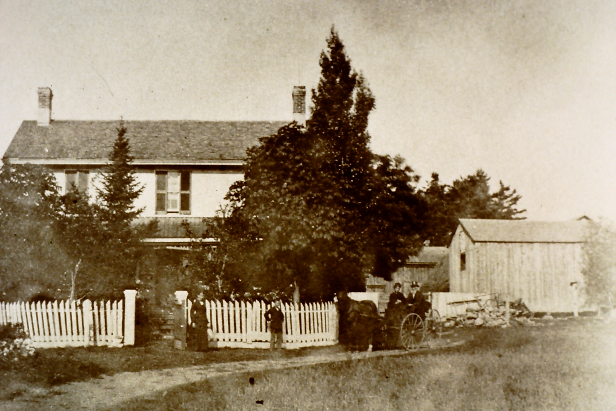 A black and white photo of a house with a white picket fence. A couple in a horse-drawn carriage are out front.