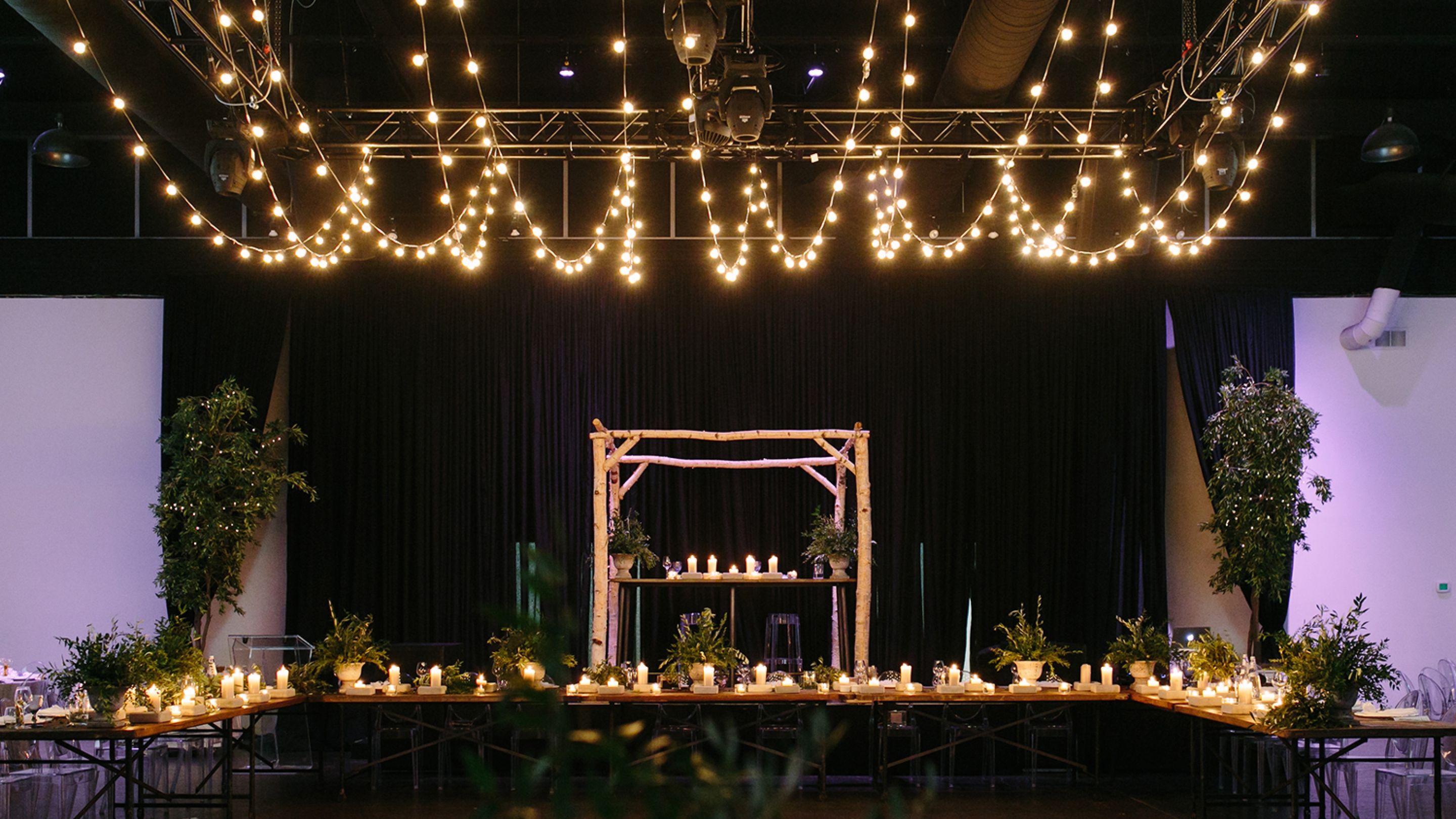 A large table set up with candles and plants in an indoor venue. Fairy lights hanging from the ceiling. 