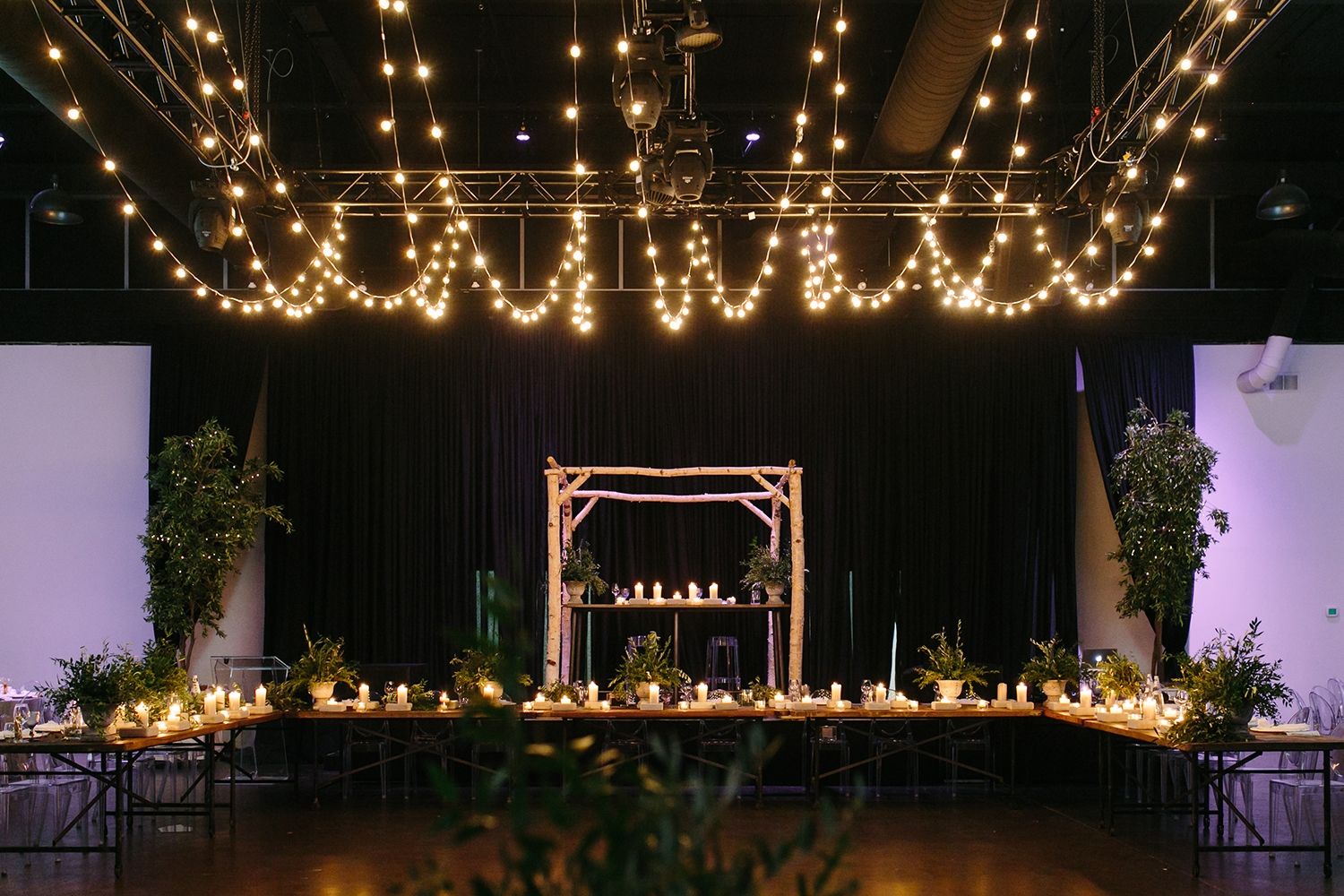 A large table set up with candles and plants in an indoor venue. Fairy lights hanging from the ceiling. 